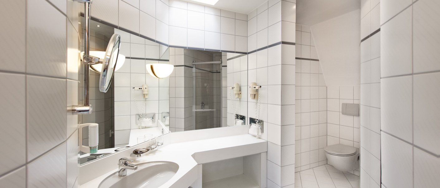 Modernes Badezimmer mit weißen Fliesen, großem Spiegel, Waschbecken und Toilette. Helle Beleuchtung und minimalistisches Design., © Hirsch Hotel