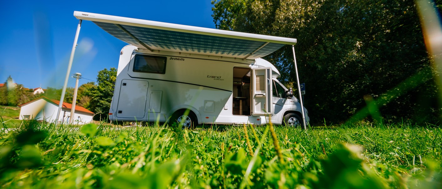 Ein Wohnmobil mit ausgefahrener Markise steht auf einer grünen Wiese unter blauem Himmel. Im Hintergrund sind Bäume und ein Gebäude zu sehen., © SMG, Thomas Niedermüller