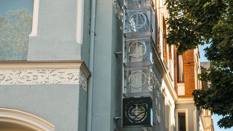 Fassade eines Gebäudes mit verzierten Elementen und einem Schild mit Logo. Ein Baum wirft Schatten auf die Wand. Klare, sonnige Atmosphäre., © SMG, Sarah Schmid