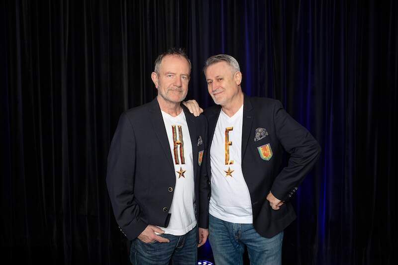 Two men in blazers and T-shirts with letters pose in front of a dark curtain., &copy; Rosenau Kultur e.V.