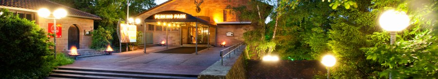 Eingang des Perkins Park in Stuttgart bei Dämmerung, umgeben von Bäumen und beleuchtet mit Laternen., © PerkinsPark