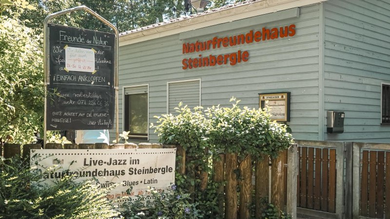 Das NaturFreundeHaus Steinbergle mit einem Schild, das Live-Jazz ankündigt, umgeben von Pflanzen und einem Holzzaun., © NaturFreundehaus Steinbergle, Stuttgart