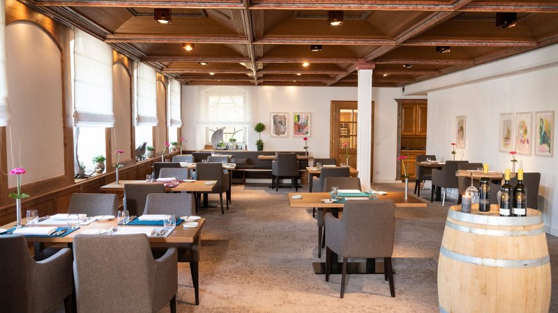 Elegantes Restaurant mit Holztischen, grauen St&uuml;hlen und einem Weinfass. Helle Fenster und moderne Kunst an den W&auml;nden schaffen eine einladende Atmosph&auml;re., &copy; Schlosshotel Monrepos