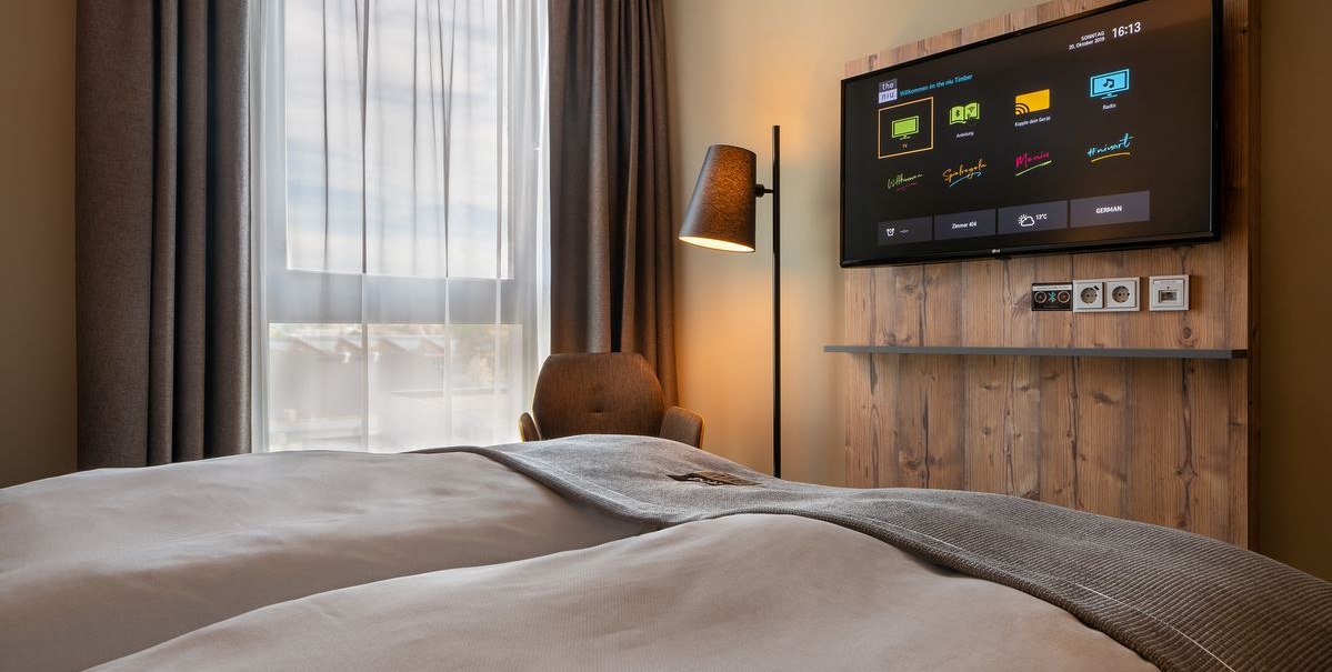 Modernes Hotelzimmer mit Doppelbett, großem Fenster, Fernseher an Holzwand und Stehlampe., © Novum Hospitality