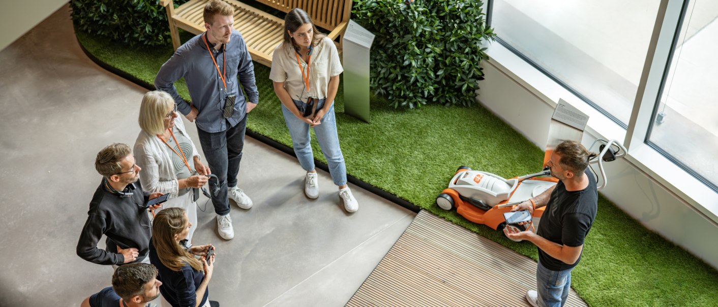 A group is listening to a presentation about a STIHL product in a modern showroom. A lawn mower can be seen in the background., © STIHL Markenwelt A group is listening to a presentation about a STIHL product in a modern showroom. A lawn mower can be seen in the background., © STIHL Markenwelt