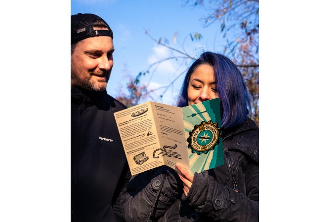 Zwei Personen im Freien, eine mit blauen Haaren liest eine Broschüre mit Kompass-Logo. Der Himmel ist blau und sonnig., © Cool-Tours StattReisen