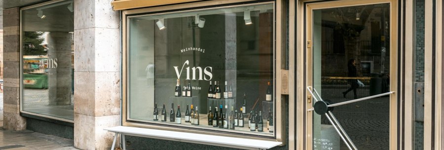 Schaufenster des Weinhandels 'Vins Gute Weine' mit Weinflaschen im Display und einer Bank davor., © SMG, Sarah Schmid