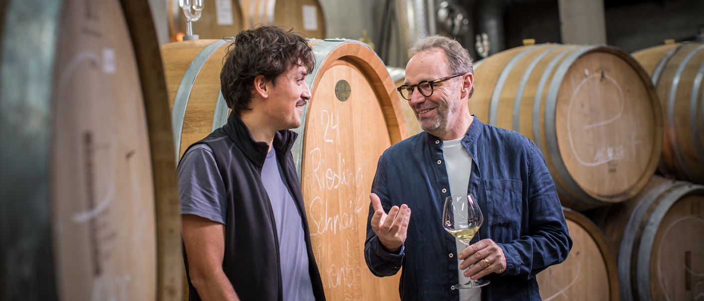 Zwei Männer stehen vor großen Holzfässern in einem Weinkeller und unterhalten sich. Einer hält ein Weinglas in der Hand., © Weingut Schnaitmann Zwei Männer stehen vor großen Holzfässern in einem Weinkeller und unterhalten sich. Einer hält ein Weinglas in der Hand., © Weingut Schnaitmann