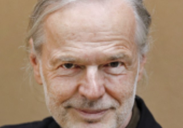 Ein älterer Mann mit grauem Haar und Bart lächelt in die Kamera. Er trägt einen dunklen Anzug vor einem neutralen Hintergrund., © Theaterhaus Stuttgart e.V.