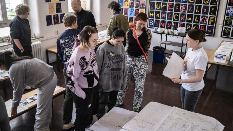 Menschen in einem Raum betrachten Zeichnungen auf Tischen. An den W&auml;nden h&auml;ngen bunte Kunstwerke. Die Atmosph&auml;re wirkt kreativ und konzentriert., &copy; Merz Akademie