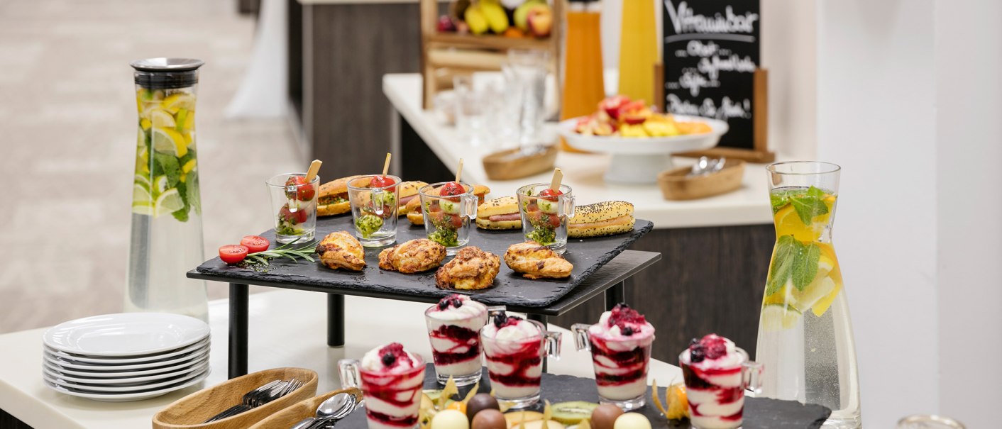 Ein Buffet mit verschiedenen Snacks, Desserts und Getränken. Im Vordergrund stehen Gläser mit Wasser und Zitronenscheiben, daneben Teller und Besteck., © ARCOTEL Hotels