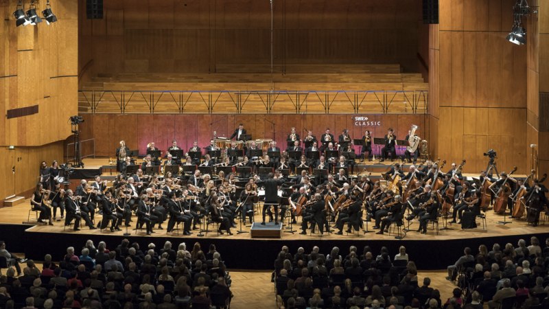 Das SWR Symphonieorchester spielt auf einer gro&szlig;en B&uuml;hne vor Publikum. Die Musiker tragen formelle Kleidung, und der Dirigent steht in der Mitte., &copy; SWR Alexander Kluge