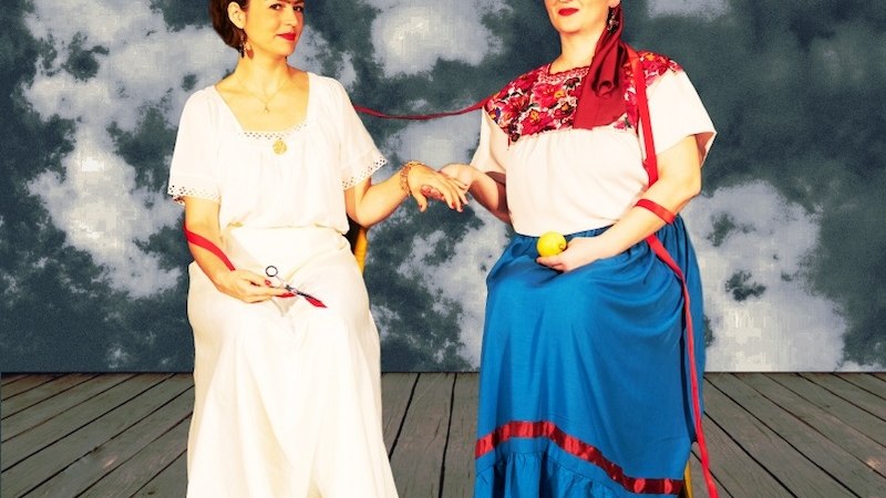 Two women in traditional dress are sitting on a wooden floor. They are connected by a red ribbon. The background shows a cloudy sky., &copy; tri-b&uuml;hne e.V.