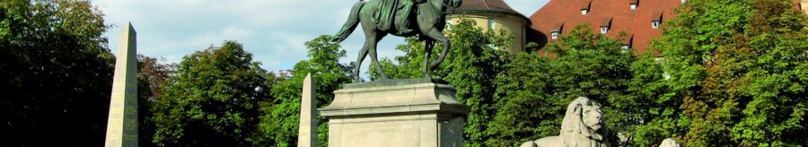 Reiterstatue auf dem Karlsplatz in Stuttgart, flankiert von Löwenstatuen und Obelisken, umgeben von Bäumen und einem historischen Gebäude im Hintergrund., © Stuttgart-Marketing GmbH