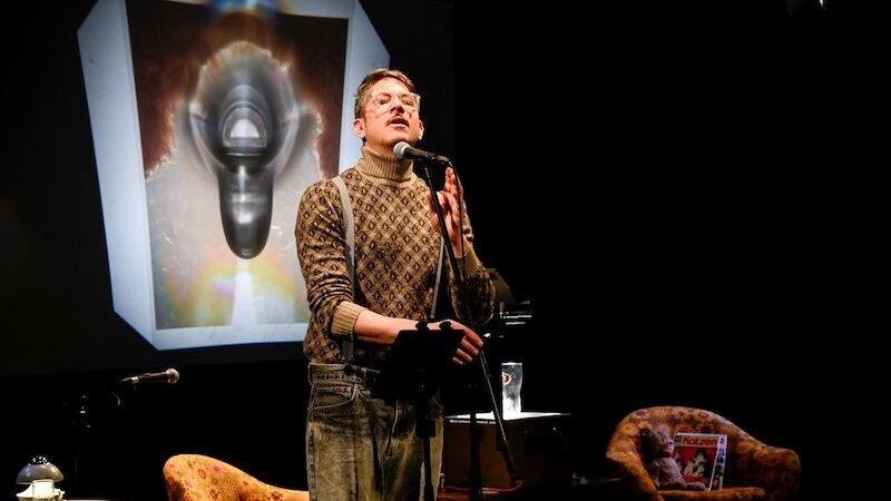 Ein Mann in weißem Hemd und Hosenträgern zeigt auf ein Foto. Im Hintergrund sind bunte Lichter und ein Kleiderständer sichtbar., © Theaterhaus Stuttgart e.V. Ein Mann in weißem Hemd und Hosenträgern zeigt auf ein Foto. Im Hintergrund sind bunte Lichter und ein Kleiderständer sichtbar., © Theaterhaus Stuttgart e.V.