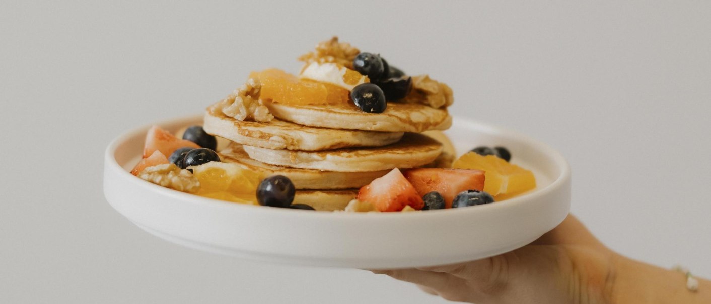A plate of pancakes, garnished with berries, walnuts and pieces of fruit, is presented by one hand., © sina fotografie, Café Lesbar, Stuttgart A plate of pancakes, garnished with berries, walnuts and pieces of fruit, is presented by one hand., © sina fotografie, Café Lesbar, Stuttgart