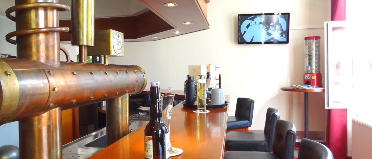Eine Bar mit hölzerner Theke, Barhockern und einem Fernseher an der Wand. Ein auffälliger Zapfhahn aus Metall ist zu sehen., © Hotel Arcis