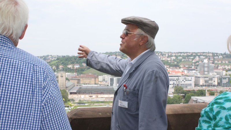 Ein älterer Mann in Arbeitskleidung und Mütze zeigt auf eine Stadtlandschaft. Im Hintergrund sind Gebäude und Hügel zu sehen., © Stuttgart-Marketing GmbH