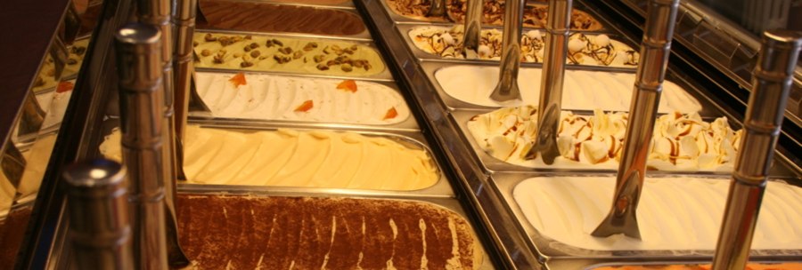 An ice cream counter with various flavors in metal containers, including chocolate, pistachio and vanilla. Metal spoons are placed in the containers., © Old Bridge, Stuttgart