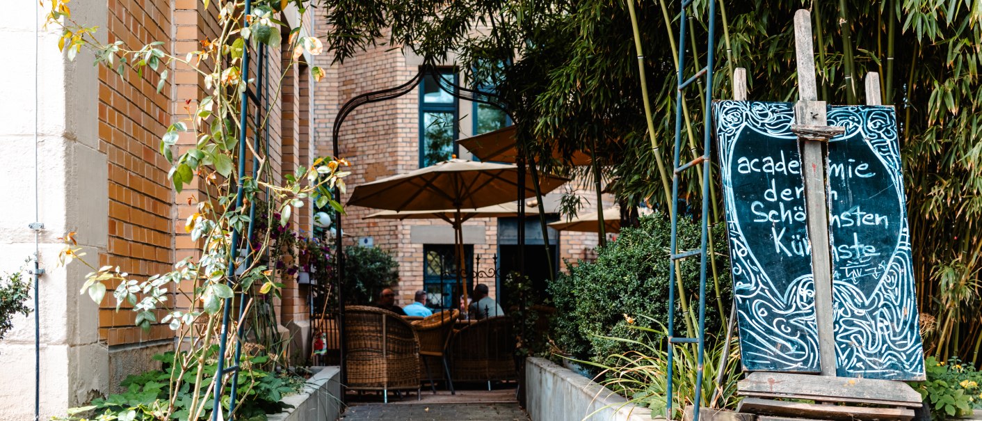 Eingang zu einem Café mit Schild 'Academie der schönsten Künste', umgeben von Pflanzen und einem Sonnenschirm., © SMG_Sarah Schmid