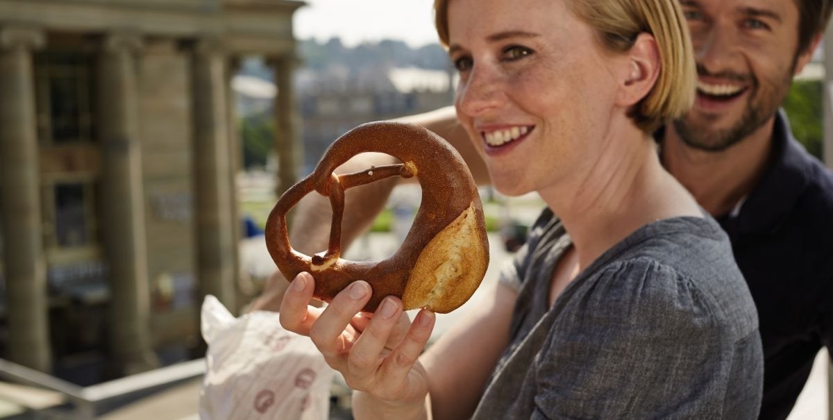 Frau hält eine schwäbische Brezel, lächelnder Mann im Hintergrund, Gebäude im Hintergrund., © TMBW, Christoph Düpper Frau hält eine schwäbische Brezel, lächelnder Mann im Hintergrund, Gebäude im Hintergrund., © TMBW, Christoph Düpper