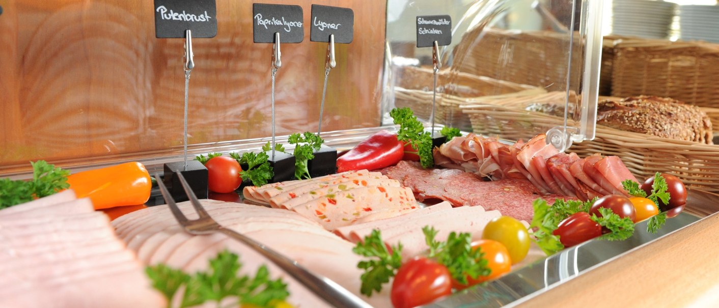Ein Buffet mit verschiedenen Wurstsorten, beschriftet mit kleinen Schildern. Dekoriert mit Tomaten und Petersilie, im Hintergrund Brotkörbe., © TOMAS
