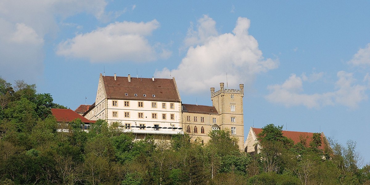 Weitenburg Castle sits enthroned on a wooded hill under a blue sky. The historic building has a striking tower and red roofs., © Hotel & Restaurant Schloss Weitenburg