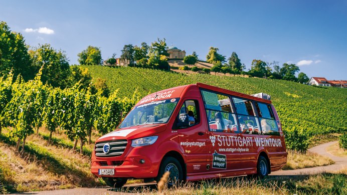 An aerial view shows the red Weintour bus of the Stuttgart Citytour driving along a paved road through a vineyard. The vine leaves are lush green, and the scene is bathed in sunlight., © SMG, Thomas Niedermüller 