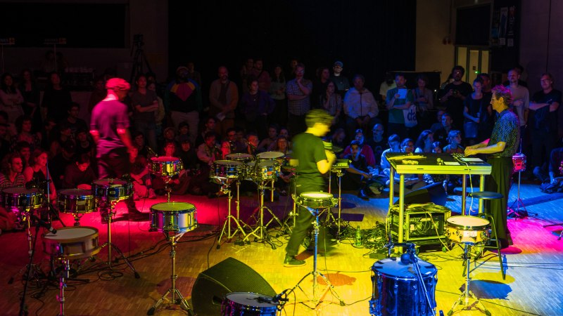 Ein Konzert mit Schlagzeug und elektronischen Instrumenten, umgeben von einer bunten Menschenmenge. Die B&uuml;hne ist in lebhaften Farben beleuchtet., &copy; IMD/Kristof Lemp