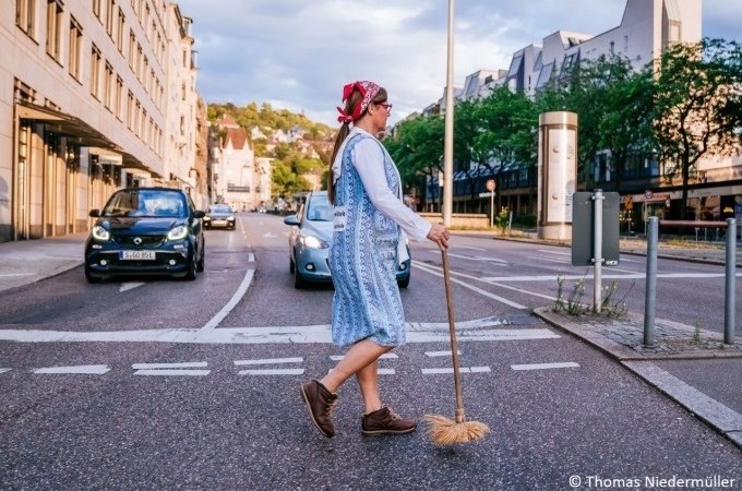 Eine Frau in traditioneller Kleidung überquert eine Straße mit einem Besen. Im Hintergrund sind Autos und Gebäude zu sehen., © Stuttgart Marketing GmbH Eine Frau in traditioneller Kleidung überquert eine Straße mit einem Besen. Im Hintergrund sind Autos und Gebäude zu sehen., © Stuttgart Marketing GmbH