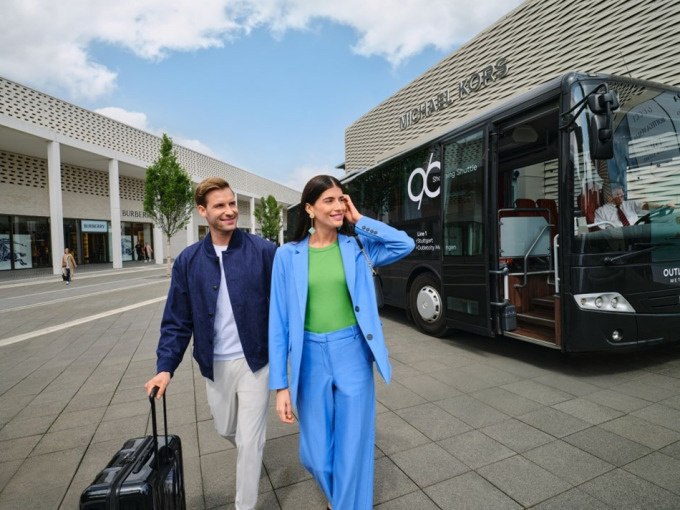 Ein Paar mit Gepäck läuft vor einem Shuttlebus in einem Einkaufszentrum. Im Hintergrund sind Geschäfte wie Michael Kors und Burberry zu sehen., © Schlienz-Tours GmbH & Co.KG Ein Paar mit Gepäck läuft vor einem Shuttlebus in einem Einkaufszentrum. Im Hintergrund sind Geschäfte wie Michael Kors und Burberry zu sehen., © Schlienz-Tours GmbH & Co.KG