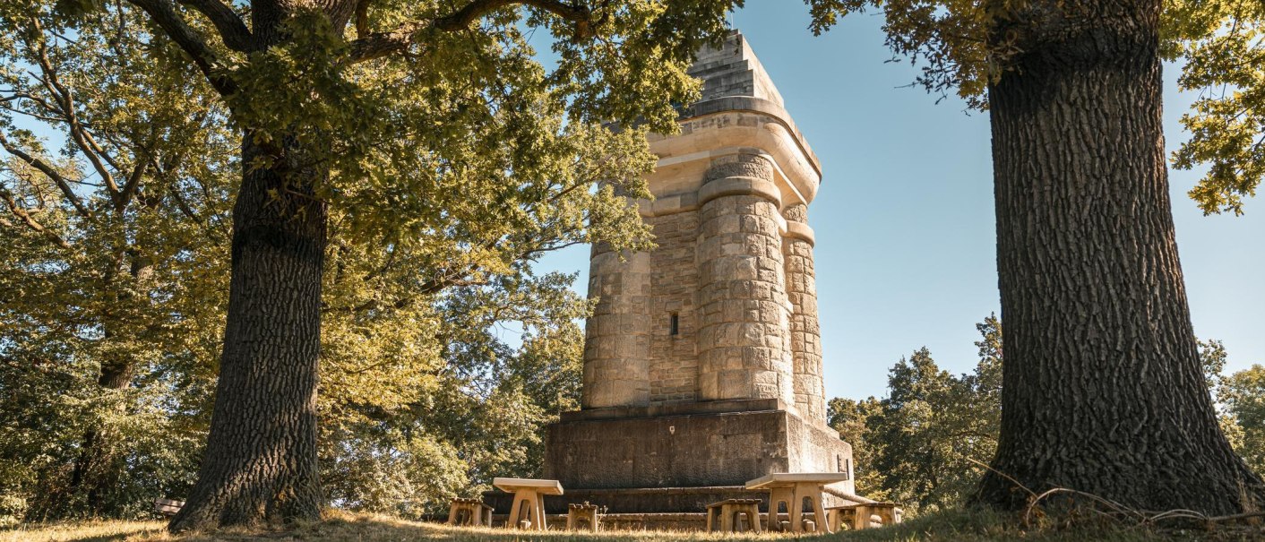 Ein steinerner Bismarckturm steht zwischen großen Bäumen. Im Vordergrund sind Bänke und Tische zu sehen. Die Sonne scheint durch das Blätterdach., © Stuttgart Marketing GmbH, Sarah Schmid