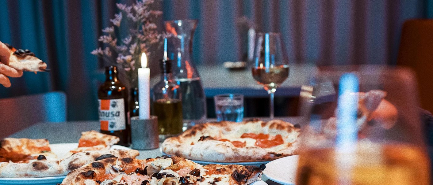 Ein Tisch in einem Restaurant mit mehreren Pizzen, Getränken und einer brennenden Kerze. Im Hintergrund sind Gläser und eine Flasche zu sehen., © Italo Disco, Stuttgart