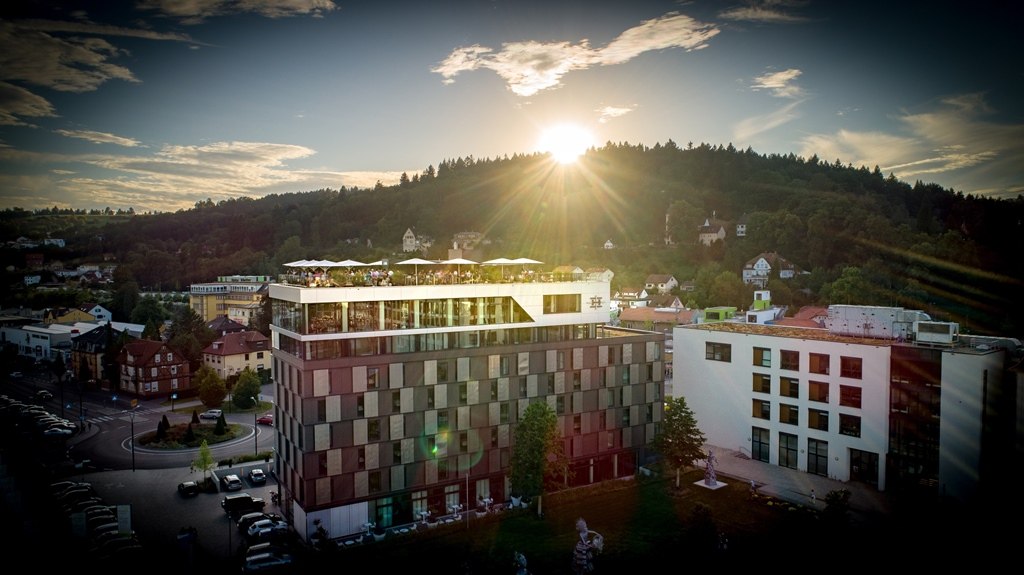 Modern hotel building at sunset, surrounded by green countryside and houses. The sun shines over a wooded hill., &copy; Hotel am Remspark