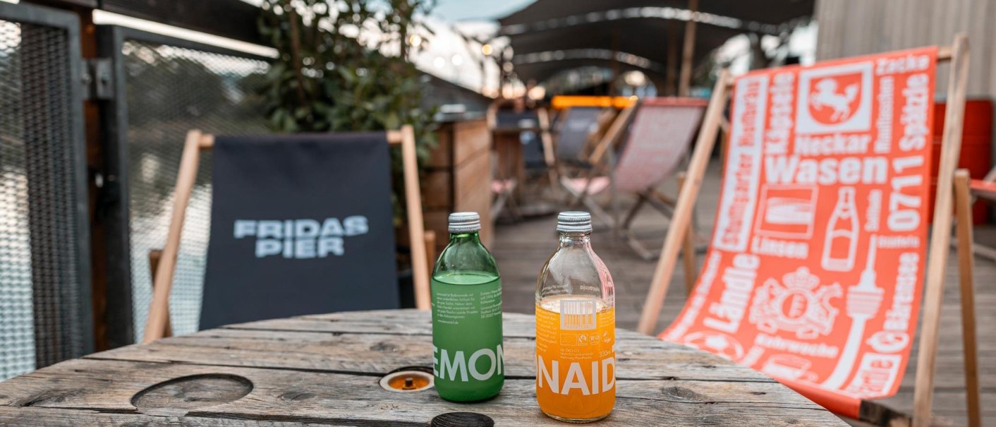 Zwei Limonadenflaschen stehen auf einem Holztisch. Im Hintergrund sind Liegest&uuml;hle mit Aufschriften bei Fridas Pier zu sehen., &copy; Stuttgart-Marketing GmbH, Sarah Schmid