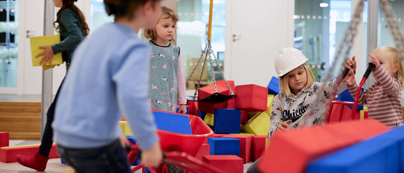 Kinder mit Bauhelmen spielen auf einer Indoor-Baustelle mit bunten Baukl&ouml;tzen und Schubkarren., &copy; Julia Ochs