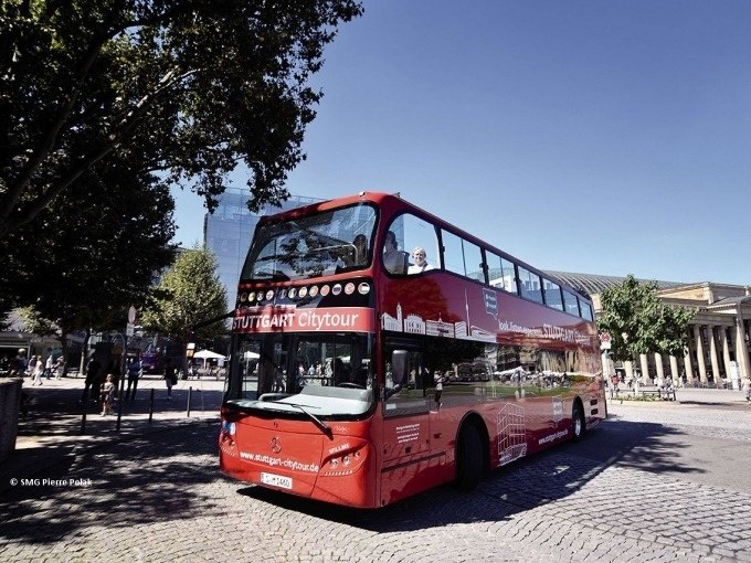 Roter Doppeldeckerbus der Stuttgart Citytour auf einem gepflasterten Platz, umgeben von Bäumen und Gebäuden., © SMG, Pierre Polak