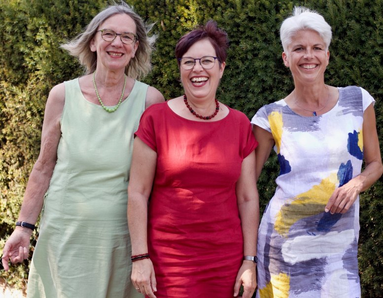 Drei Frauen in bunten Sommerkleidern stehen lächelnd vor einer grünen Hecke., © Ursel Krieg