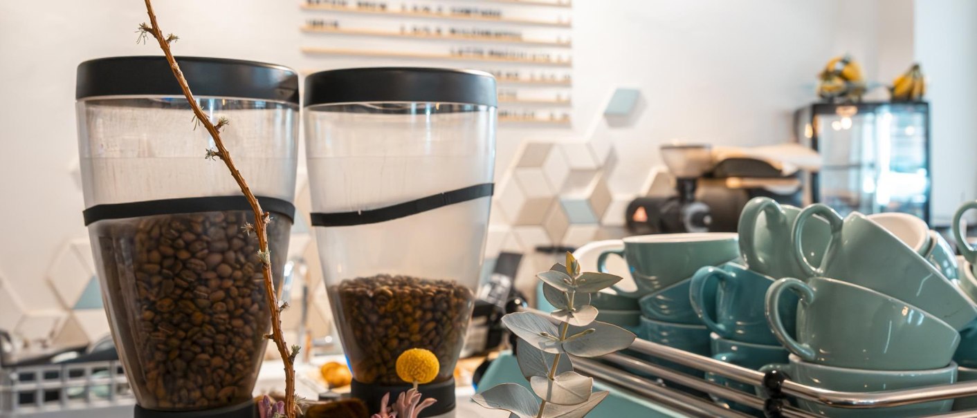 Kaffeebohnenbehälter und türkisfarbene Tassen in einem Café. Im Hintergrund sind geometrische Wanddekorationen und ein Kühlschrank zu sehen., © Stuttgart-Marketing GmbH, Sarah Schmid