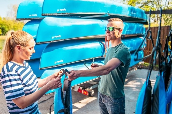 Two people are standing in front of a pile of blue canoes. They are holding life jackets and preparing for a canoe trip., © Cool-Tours StattReisen Two people are standing in front of a pile of blue canoes. They are holding life jackets and preparing for a canoe trip., © Cool-Tours StattReisen