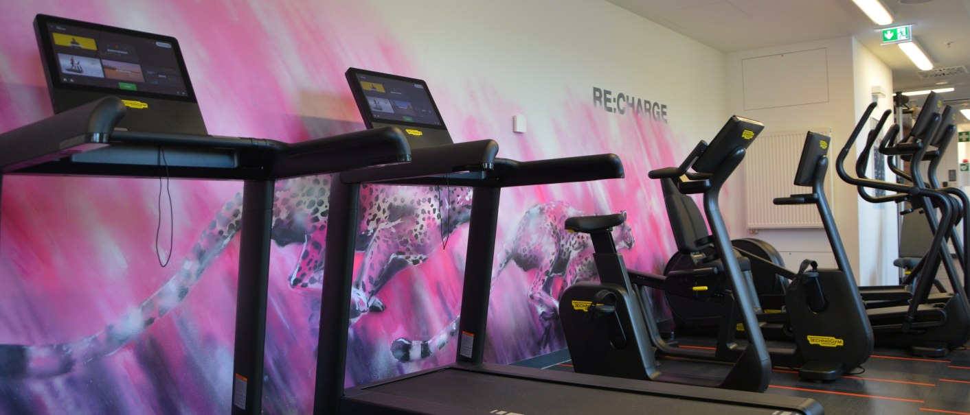 Ein Fitnessstudio mit modernen Laufbändern und Crosstrainern vor einer rosa Wand mit einem Gepardenmotiv. Der Raum wirkt einladend und motivierend., © Aloft Stuttgart Ein Fitnessstudio mit modernen Laufbändern und Crosstrainern vor einer rosa Wand mit einem Gepardenmotiv. Der Raum wirkt einladend und motivierend., © Aloft Stuttgart