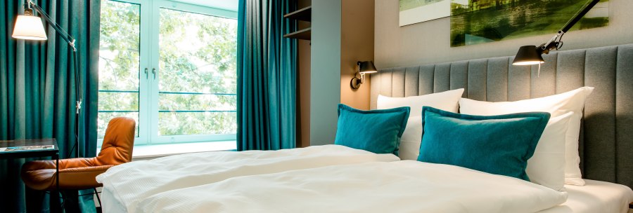 Modernes Hotelzimmer mit gro&szlig;em Bett, blauen Kissen und Vorh&auml;ngen. Ein Fenster bietet Blick ins Gr&uuml;ne. Ein Schreibtisch mit Stuhl steht daneben., &copy; Motel One - Stuttgart Hauptbahnhof