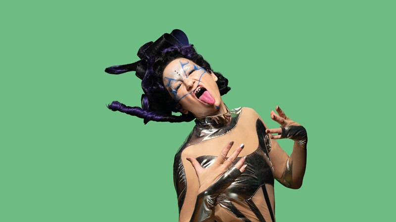 Eine Person in einem futuristischen Outfit mit auffälligem Make-up posiert vor einem grünen Hintergrund und streckt die Zunge heraus., © Württembergische Staatstheater Stuttgart