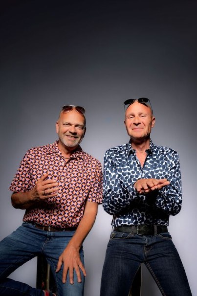 Two men in patterned shirts and jeans sit next to each other. Both wear sunglasses on their heads and smile against a gray background., &copy; Theaterhaus Stuttgart e.V.