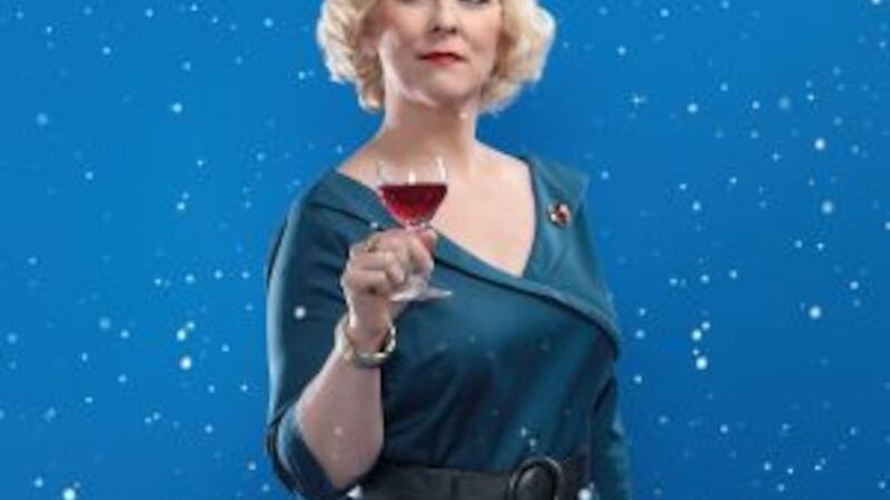 Eine Frau in einem blauen Kleid hält ein Weinglas vor einem blauen Hintergrund mit Schneeflocken., © Altes Schauspielhaus und Komödie im Marquardt e.V. Eine Frau in einem blauen Kleid hält ein Weinglas vor einem blauen Hintergrund mit Schneeflocken., © Altes Schauspielhaus und Komödie im Marquardt e.V.