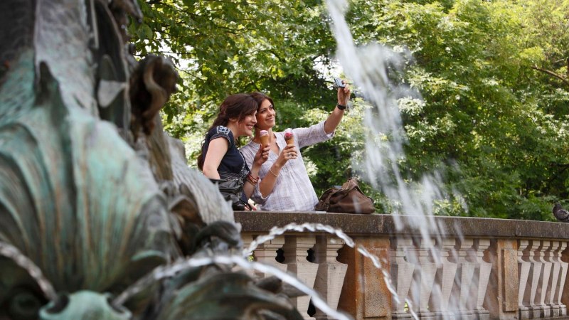 Zwei Frauen machen ein Selfie am Galateabrunnen, halten Eis in der Hand. Im Hintergrund sind Bäume und ein Wasserspritzer zu sehen., © Stuttgart-Marketing GmbH Christoph Düpper