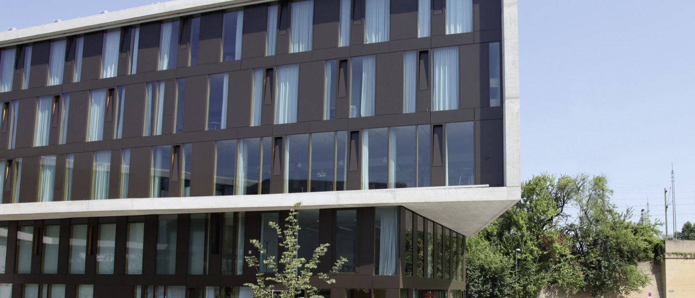 A modern building with a glass facade, a terrace, and trees in the foreground. The Stuttgart Neckarpark Youth Hostel under a clear sky., © DJH Landesverband Baden-Württemberg e.V. A modern building with a glass facade, a terrace, and trees in the foreground. The Stuttgart Neckarpark Youth Hostel under a clear sky., © DJH Landesverband Baden-Württemberg e.V.