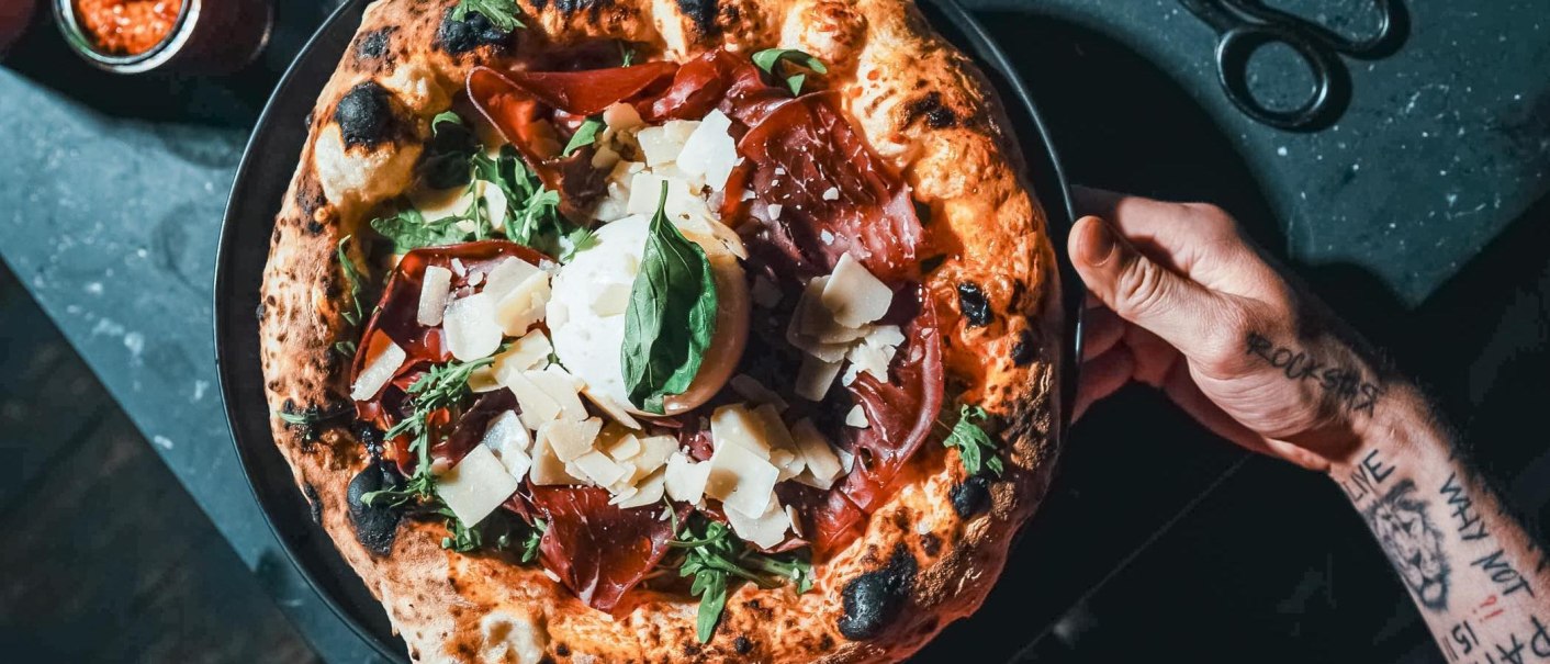 Von oben: Pizza mit luftiger Kruste, Schinken, Rucola, Käse und Basilikum auf schwarzem Teller, gehalten von einer tätowierten Hand., © Connective Media Von oben: Pizza mit luftiger Kruste, Schinken, Rucola, Käse und Basilikum auf schwarzem Teller, gehalten von einer tätowierten Hand., © Connective Media