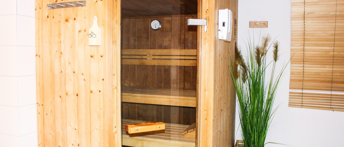 Ein Saunaraum mit Holzwänden und Glastür. Innen sind Holzbänke und eine Uhr sichtbar. Rechts steht eine Pflanze in einem Holzkasten., © TOMAS