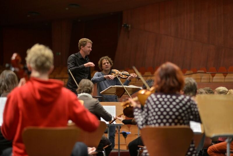 Ein Dirigent und ein Violinist leiten eine Orchesterprobe in einem Konzertsaal. Musiker sitzen auf Stühlen und spielen Instrumente., © il Gusto Barocco - Stuttgarter Barockorchester